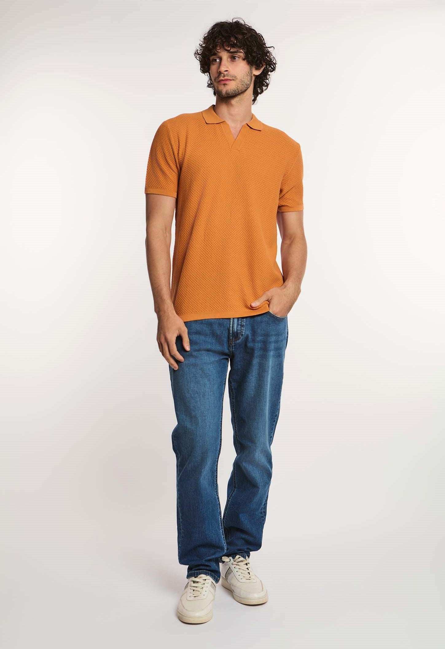 Man in an orange textured polo shirt, blue jeans, and white sneakers standing casually